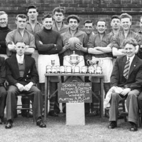 Players and officials of South Hetton Rovers AFC (or possibly South Hetton Colliery Welfare AFC) after the team had won both the Division 2 Championship of the Houghton & District League and the Murton A M Cup in the season 1955/6.  My father, John Sheldon Wesley Clark is seated at the extreme left of the picture, next to a Mr Beresford, I believe.  Although the faces of most of the rest of the officials are familiar, the only other one I can name is Sammy Mitford, who is standing towards the right, wearing an overcoat and scarf.  I think the player standing third from the right was called Whittaker and came from Hetton Downs.  I have no knowledge of the other men's identities.  The shot seems to have been taken at the Colliery Welfare Ground, behind Hawthorn Terrace (as it then was), near where the cricket club now has its clubhouse.                    .Photograph and caption from Sheldon Clark.